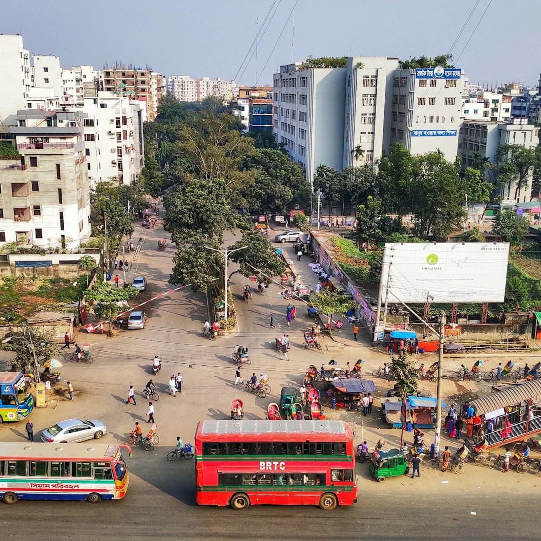 A wide illustration of Dhaka at golden hour: rickshaws moving along a wide boulevard, the National Parliament building visible through palm trees, and a warm amber sky above.