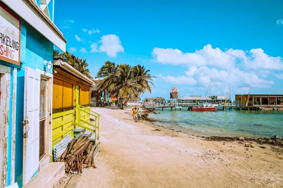 A sunlit view of the Swing Bridge over Haulover Creek in Belize City, with wooden storefronts and palm trees lining the waterfront under a bright Caribbean sky.