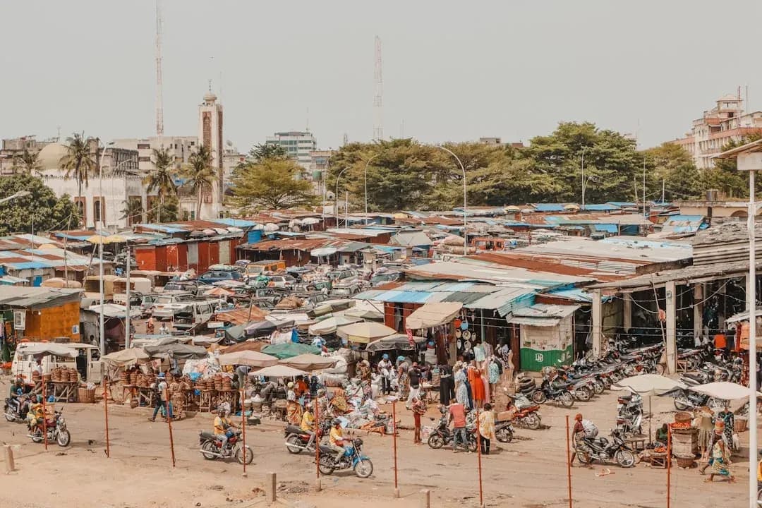 A freelance contractor in Cotonou working at a desk with invoices and a laptop, with the lagoon and the market stalls of the Dantokpa area visible through the window.