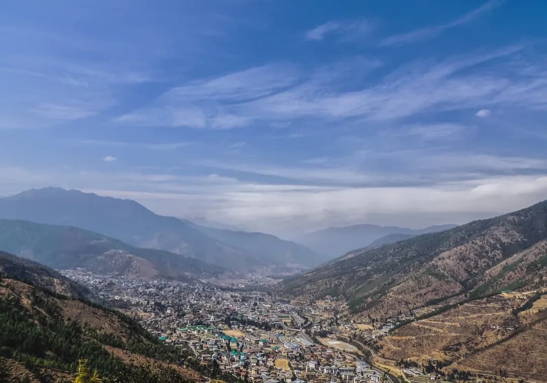 A contractor working at a wooden desk in Thimphu with the Tiger's Nest monastery visible in the distance through a window, warm afternoon light falling across the papers and laptop.