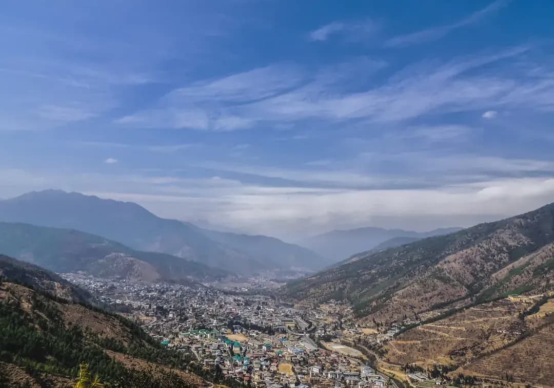 A contractor working at a wooden desk in Thimphu with the Tiger's Nest monastery visible in the distance through a window, warm afternoon light falling across the papers and laptop.