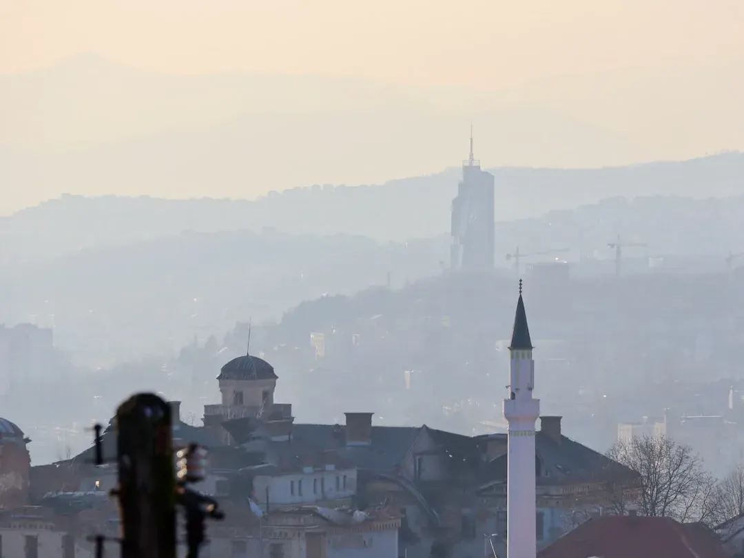 A warm illustration of Sarajevo at golden hour: the Bascarsija old town rooftops, the river Miljacka, and green hills rising behind the city under an amber sky.