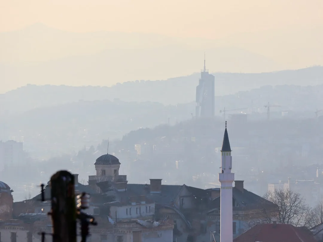 A warm illustration of Sarajevo at golden hour: the Bascarsija old town rooftops, the river Miljacka, and green hills rising behind the city under an amber sky.