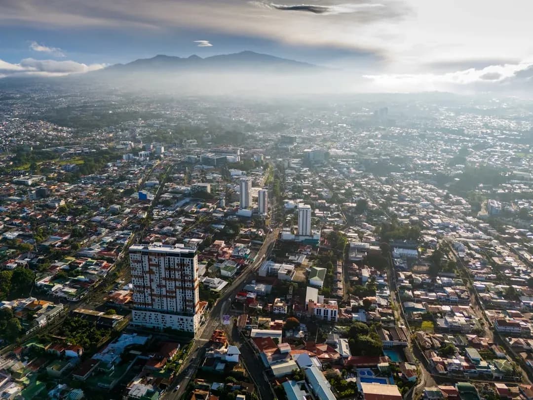 A warm wide illustration of San Jose, Costa Rica at golden hour: the National Theatre and red-roofed downtown buildings, green Central Valley mountains behind, and a clear amber sky.
