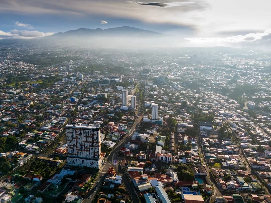 A warm wide illustration of San Jose, Costa Rica at golden hour: the National Theatre and red-roofed downtown buildings, green Central Valley mountains behind, and a clear amber sky.
