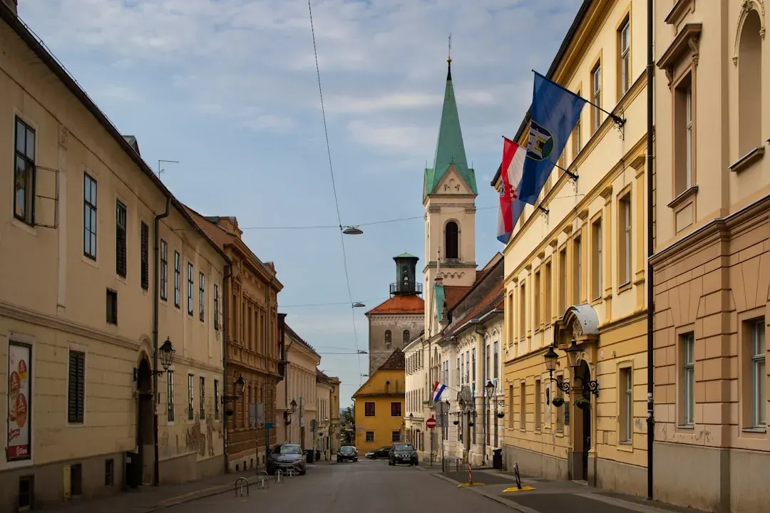 A wide warm illustration of Zagreb at golden hour: the red rooftops of the Upper Town, the twin spires of Zagreb Cathedral, and the green Ban Jelacic Square below under an amber sky.