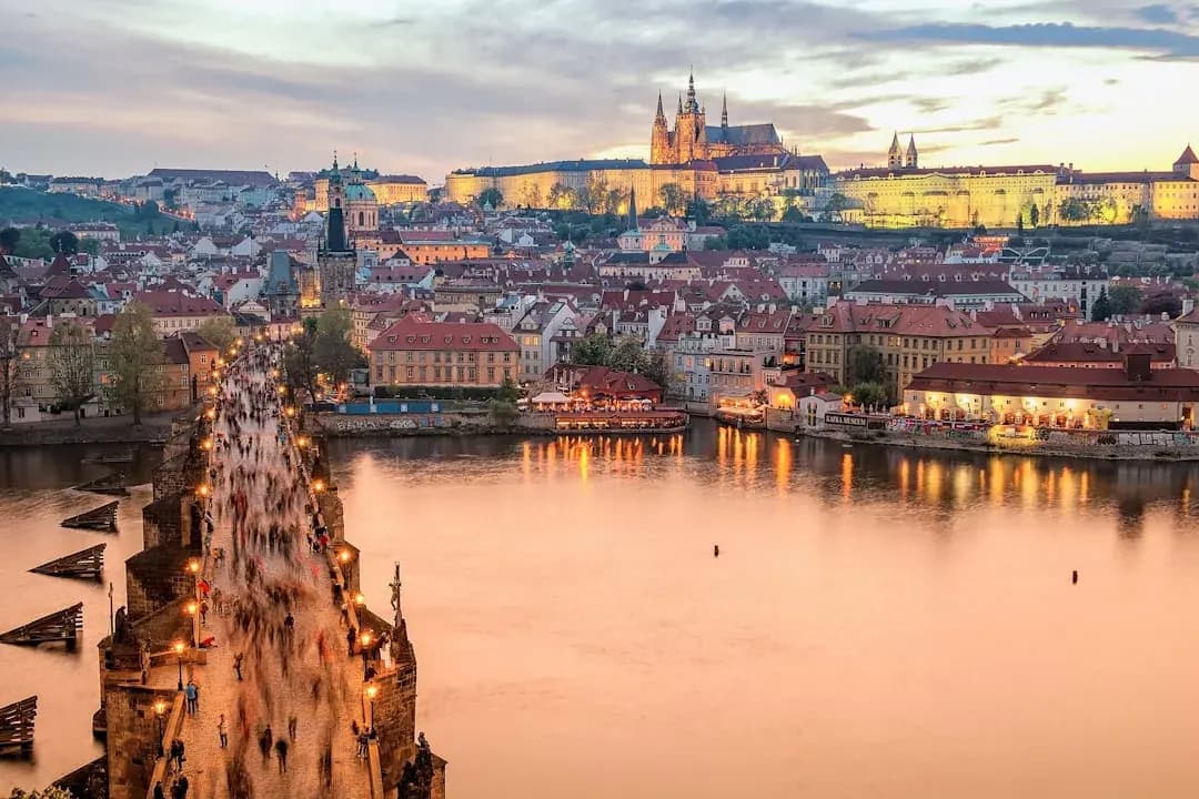 A warm wide illustration of Prague at golden hour, the Charles Bridge and its statues crossing the Vltava river toward Prague Castle and St Vitus Cathedral on the hill, amber evening light over the red rooftops of Mala Strana.