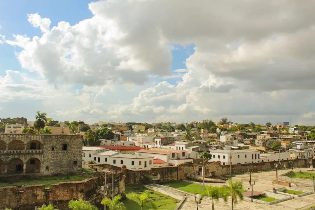 A warm illustration of Santo Domingo's colonial Zona Colonial at golden hour, with pastel Spanish-era buildings along Calle Las Damas and palm trees against a soft amber sky.