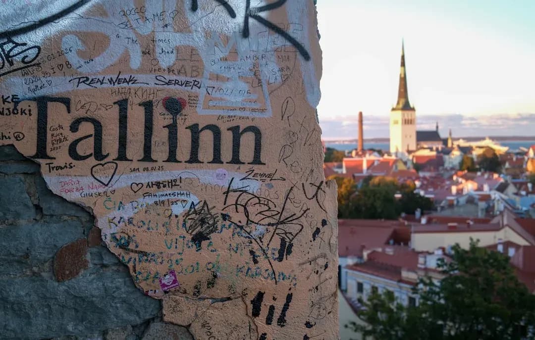 A wide warm illustration of Tallinn at golden hour: the medieval Old Town spires of St Olaf's Church and Toompea Castle above terracotta rooftops, with the glass towers of the modern business district beyond and the Baltic Sea catching amber light.
