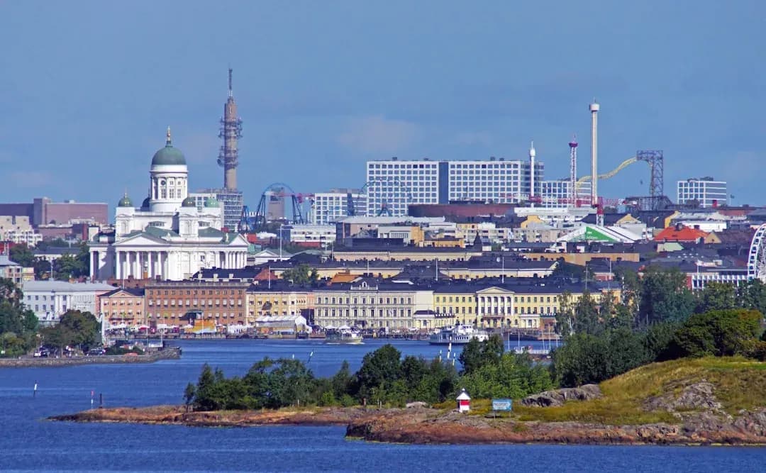 A wide warm illustration of Helsinki on a bright summer evening: the pale green domes and white columns of Helsinki Cathedral above Senate Square, the harbour and market hall to the side, and a soft amber Nordic sky.