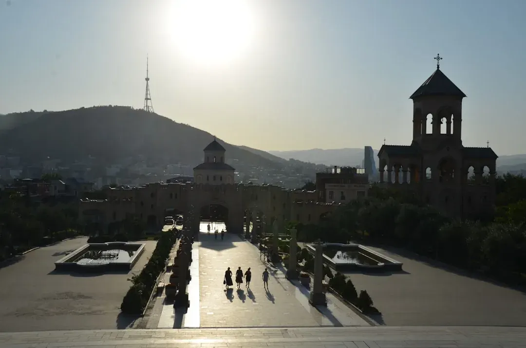 A wide warm illustration of Tbilisi at golden hour: the old town's pastel balconied houses below Narikala fortress, the Kura river curving through, and amber light on the hillside.