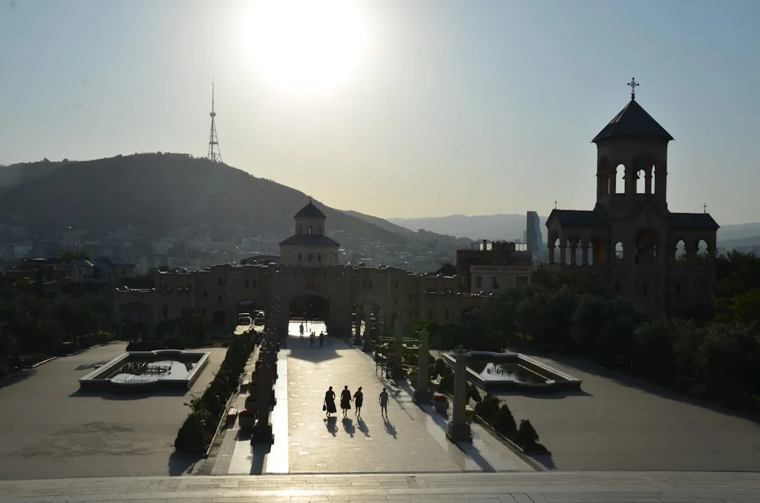 A wide warm illustration of Tbilisi at golden hour: the old town's pastel balconied houses below Narikala fortress, the Kura river curving through, and amber light on the hillside.