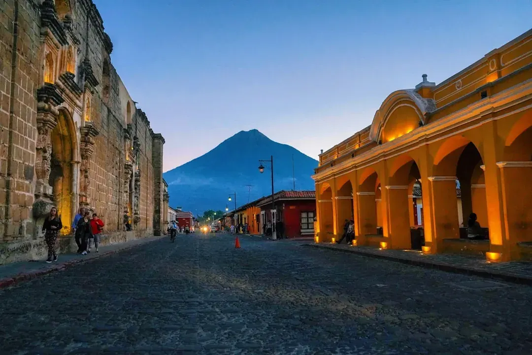 A warm illustration of Antigua Guatemala at golden hour, cobbled streets and pastel colonial facades below the Volcan de Agua, with a clear amber sky.