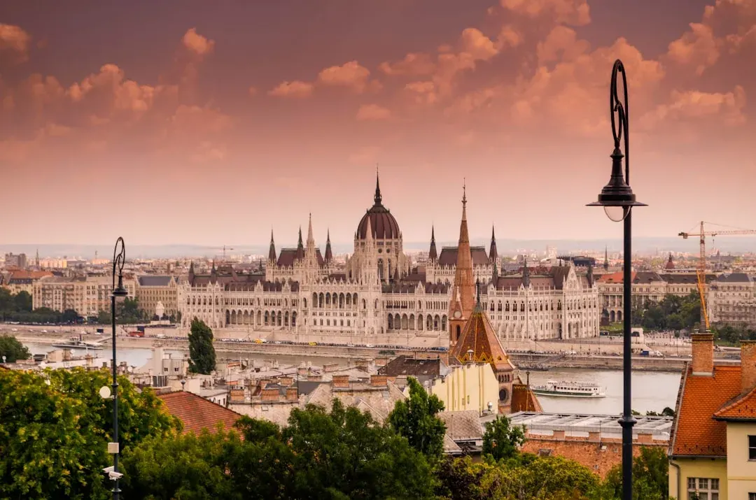 A wide warm illustration of Budapest at golden hour: the Hungarian Parliament Building along the Danube, the Chain Bridge crossing to Buda Castle on the hill, an amber sky above the river.