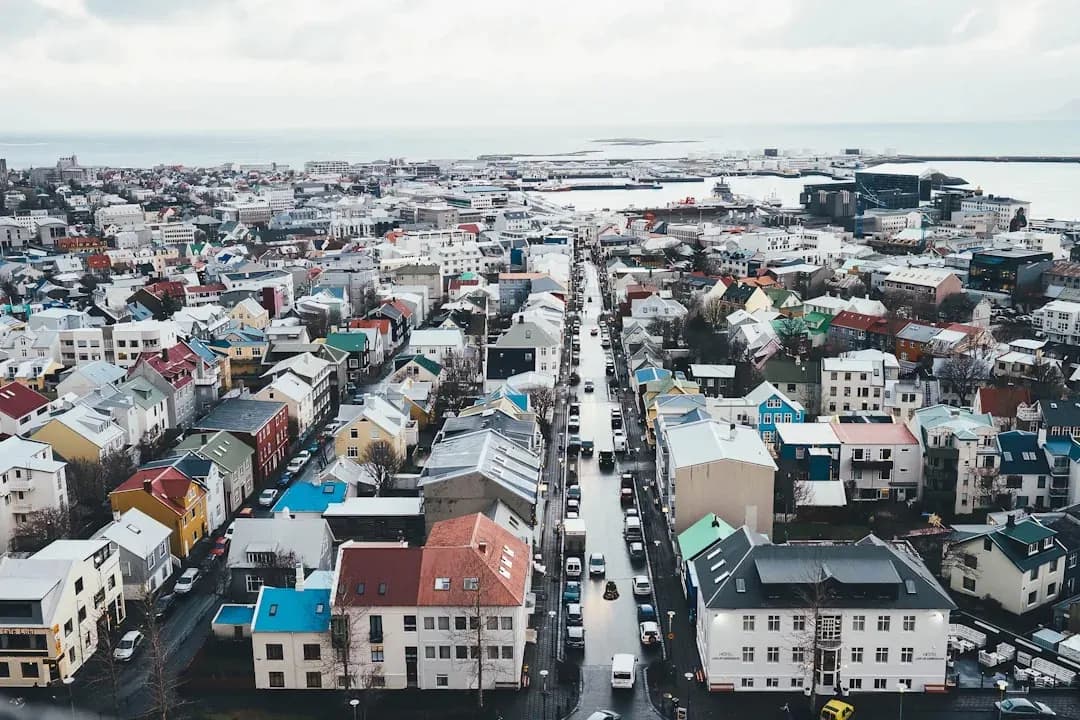 A warm illustration of Reykjavik on a clear day: the Hallgrimskirkja church tower rising above the colourful rooftops of the old harbour district, with soft mountains in the distance.