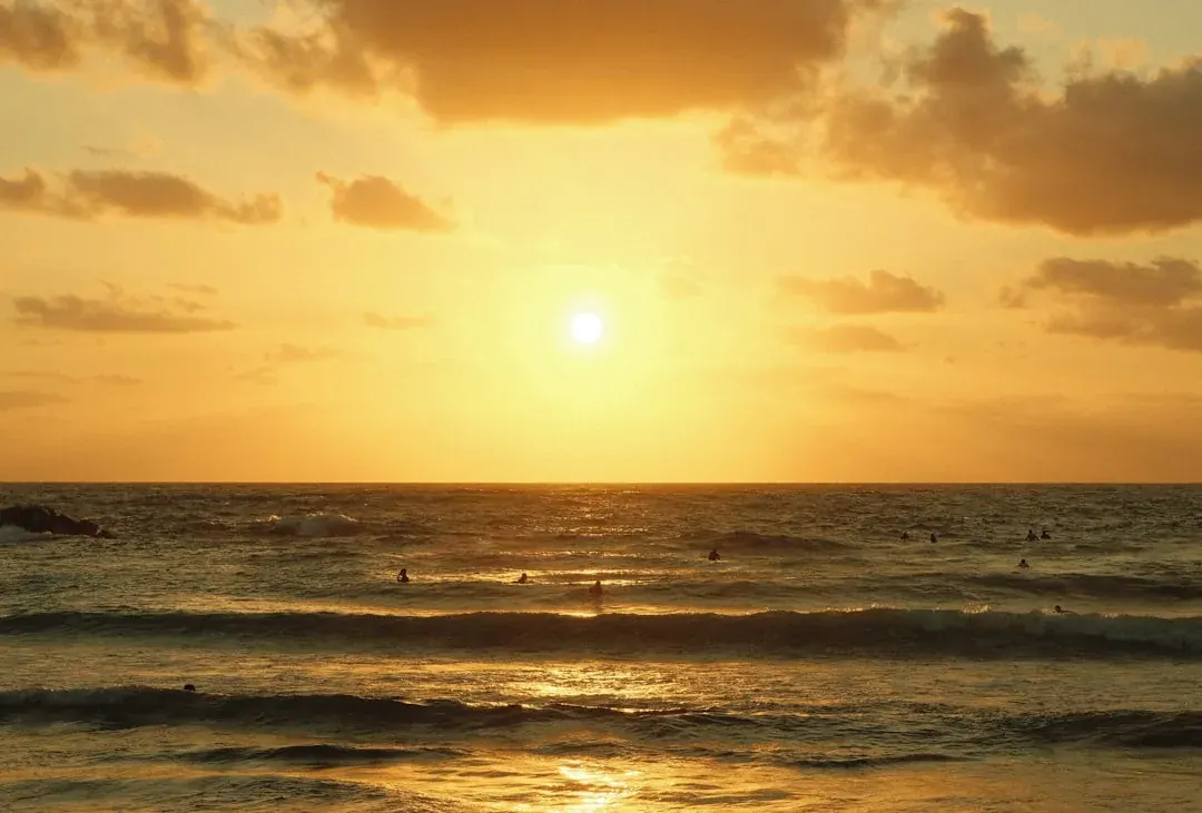 A warm wide illustration of Tel Aviv at golden hour: the Mediterranean shoreline curving past the white Bauhaus buildings of the city centre, sailboats on the water, and the towers of the business district behind under an amber sky.