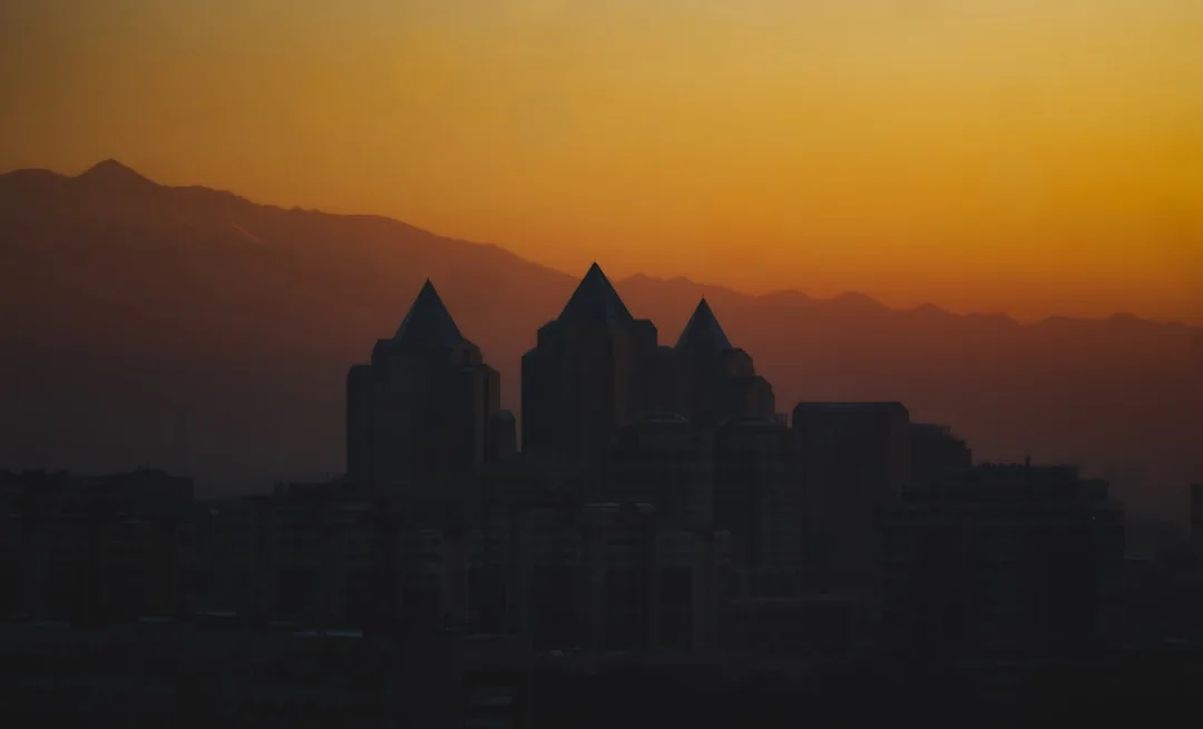 A wide warm illustration of Almaty at golden hour, with the snow-capped Trans-Ili Alatau mountains behind the city and tree-lined avenues in the foreground under an amber sky.