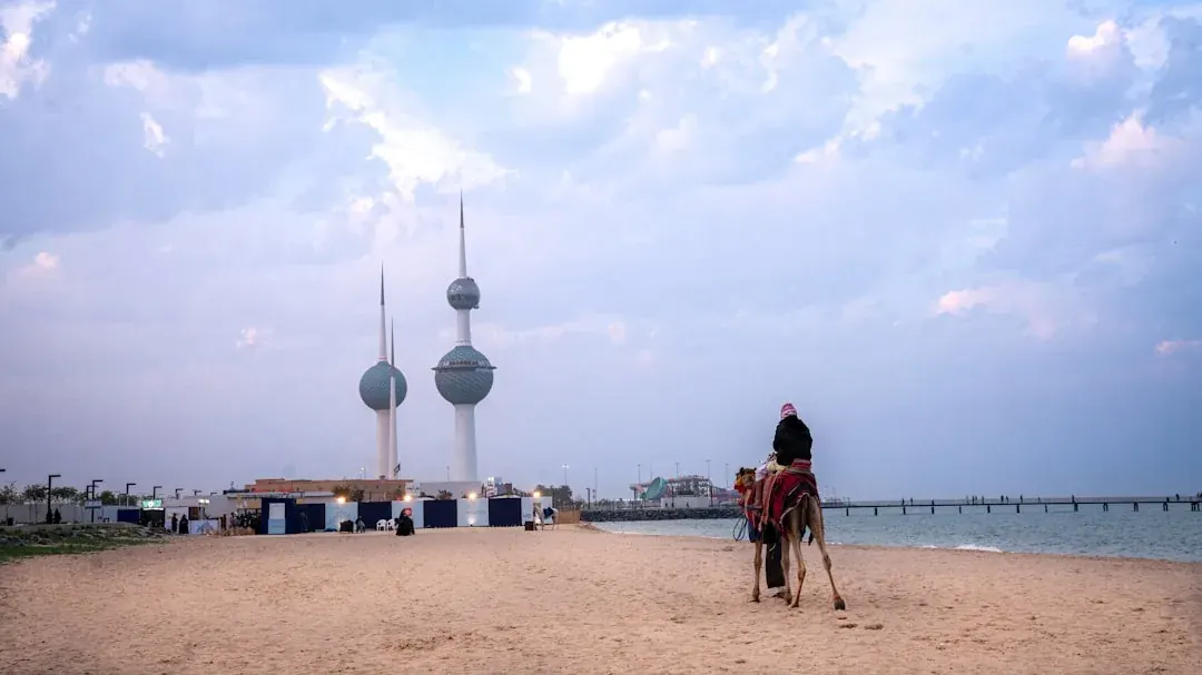 A wide warm illustration of Kuwait City at golden hour, the Kuwait Towers rising over the Arabian Gulf, dhow boats on the water, and the waterfront corniche in the foreground under a soft amber sky.