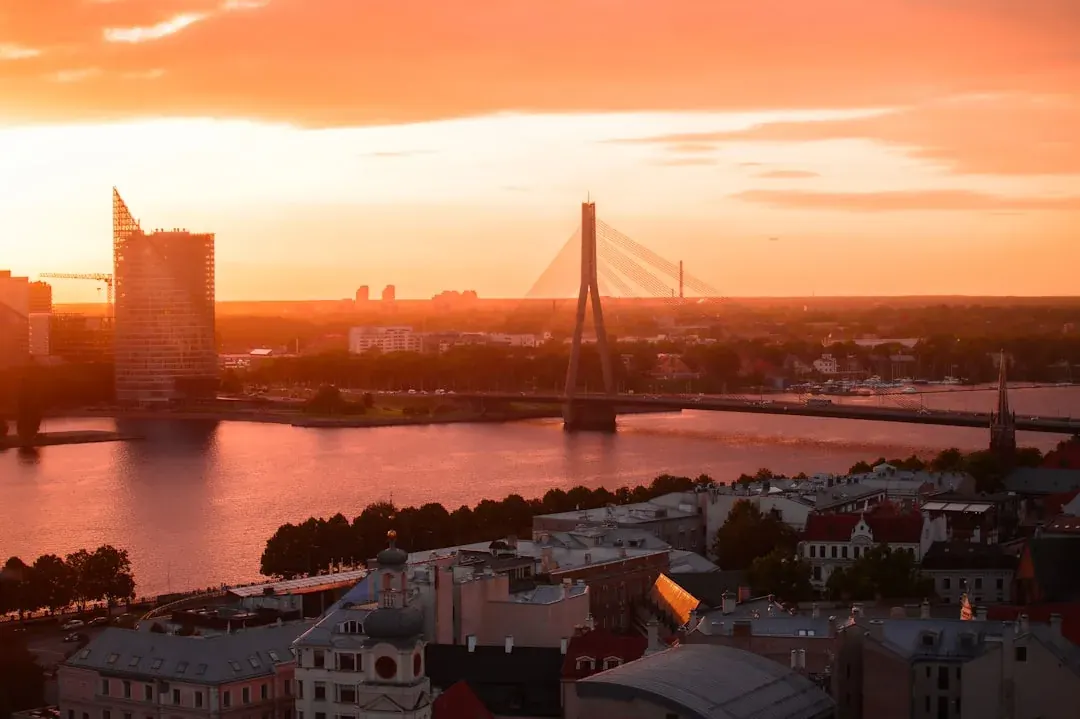A warm wide illustration of Riga at golden hour: the spire of St Peter's Church rising over the pastel facades of the Old Town, the Daugava river catching amber light, and red rooftops below a clear sky.
