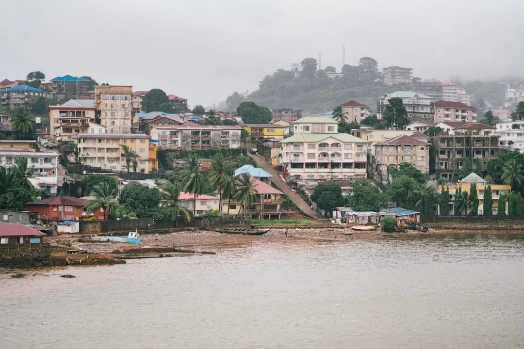 A contractor in Liberia working at a desk overlooking the streets of Monrovia, with Atlantic Ocean light through the window and papers spread across the table.