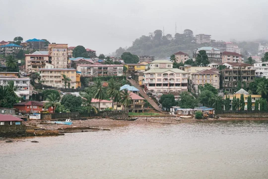 A contractor in Liberia working at a desk overlooking the streets of Monrovia, with Atlantic Ocean light through the window and papers spread across the table.