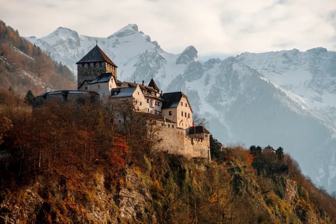 A wide view of Vaduz Castle on a wooded hillside above the Rhine valley, with the Liechtenstein capital visible below and the Alps rising in the background.