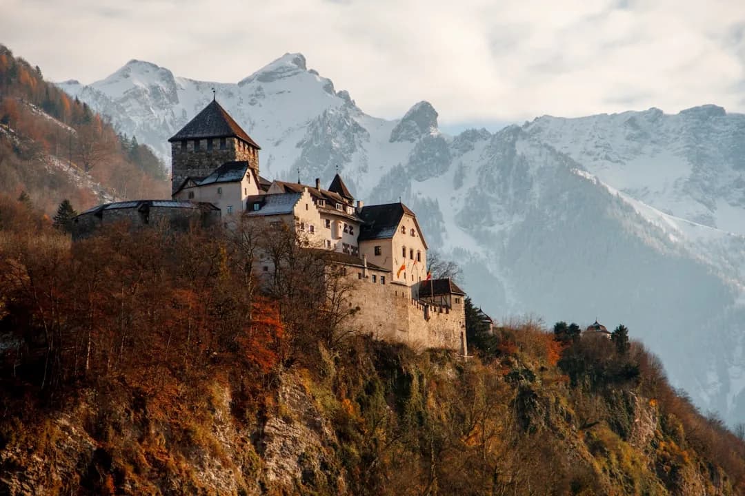 A wide view of Vaduz Castle on a wooded hillside above the Rhine valley, with the Liechtenstein capital visible below and the Alps rising in the background.