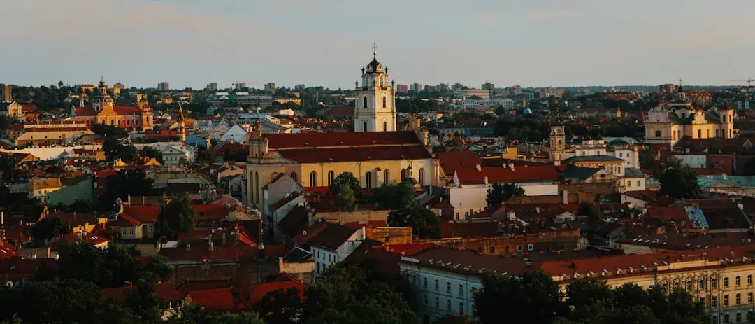 A warm illustration of Vilnius old town at golden hour, the Gediminas Tower on its green hill above terracotta rooftops, the baroque spires of the old town below, and a clear amber sky.
