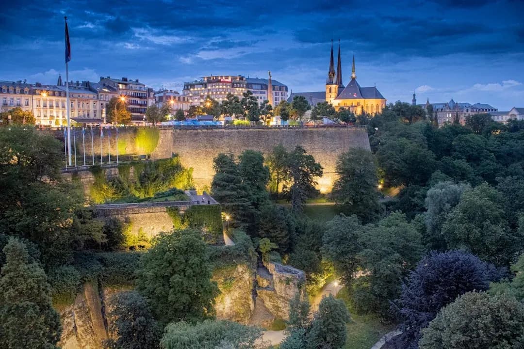 A warm wide illustration of Luxembourg City at golden hour: the Adolphe Bridge arching over the green Pétrusse valley, the old town ramparts and spires of the Ville Haute behind, a soft amber sky above.