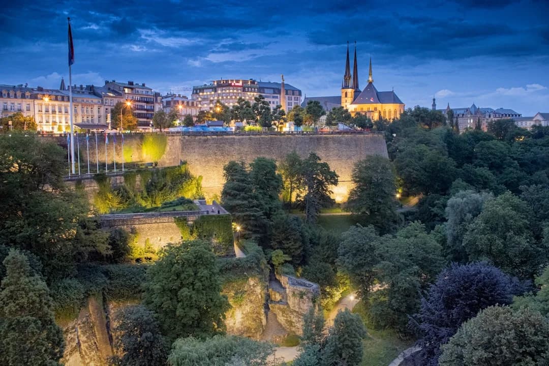 A warm wide illustration of Luxembourg City at golden hour: the Adolphe Bridge arching over the green Pétrusse valley, the old town ramparts and spires of the Ville Haute behind, a soft amber sky above.