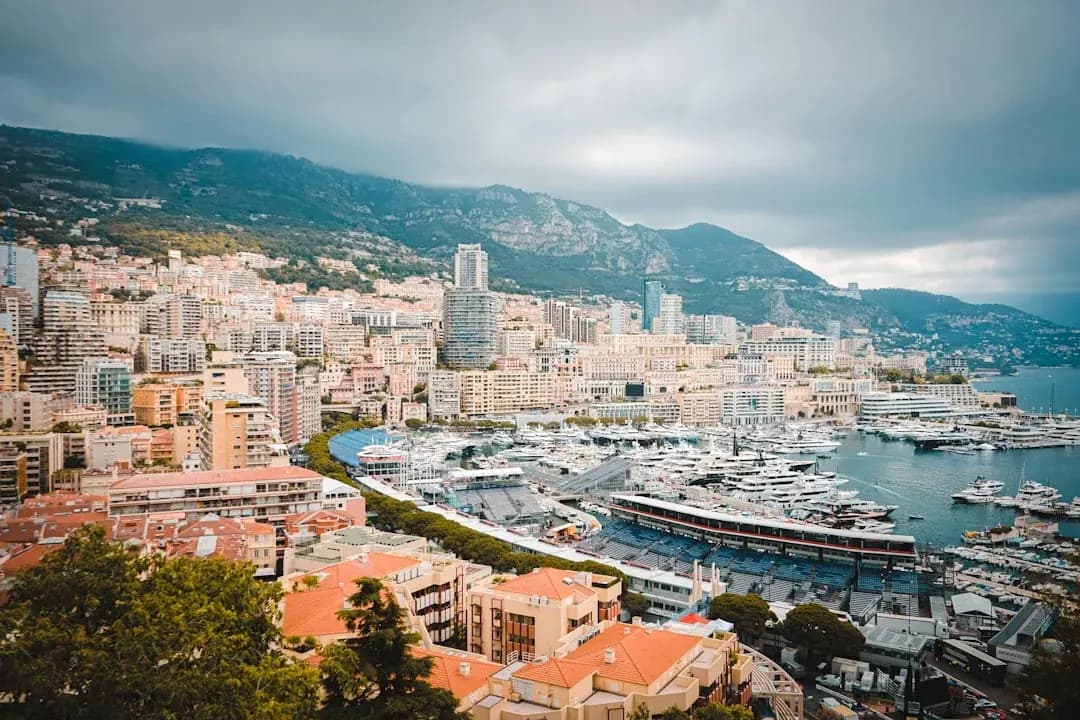 The Port Hercule harbour in Monaco at midday, with the distinctive pink Casino gardens visible on the headland and private yachts moored in the foreground.