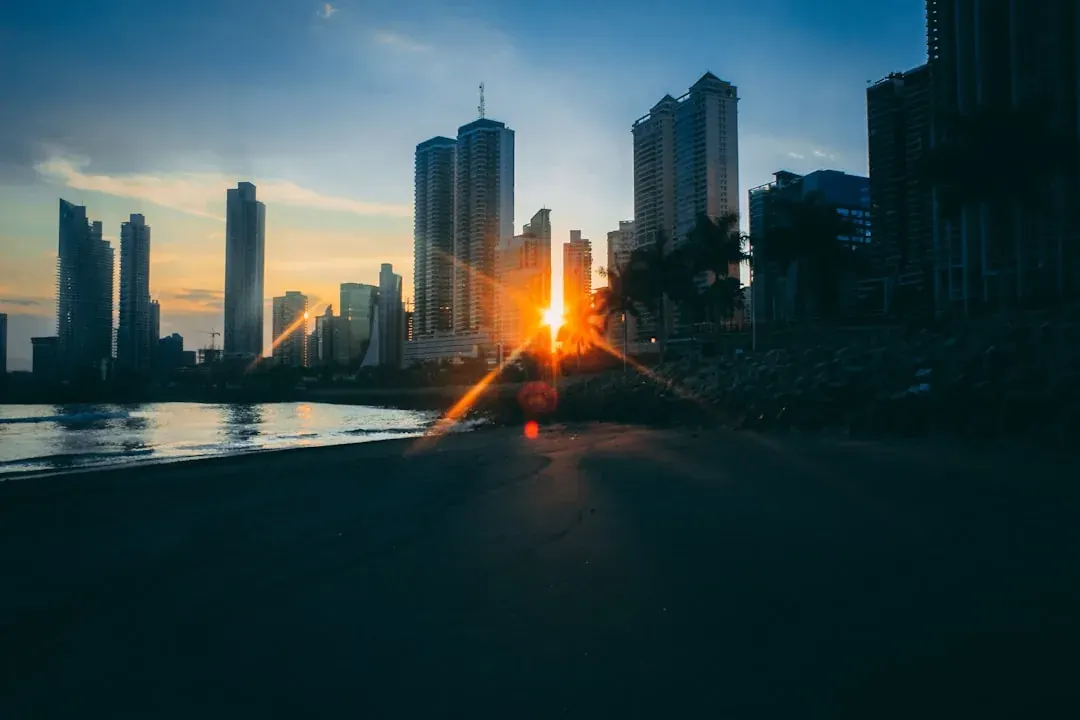 A wide warm illustration of Panama City at golden hour: the Cinta Costera waterfront curving past the modern skyline of glass towers, with ships waiting near the Pacific entrance to the Panama Canal under an amber sky.