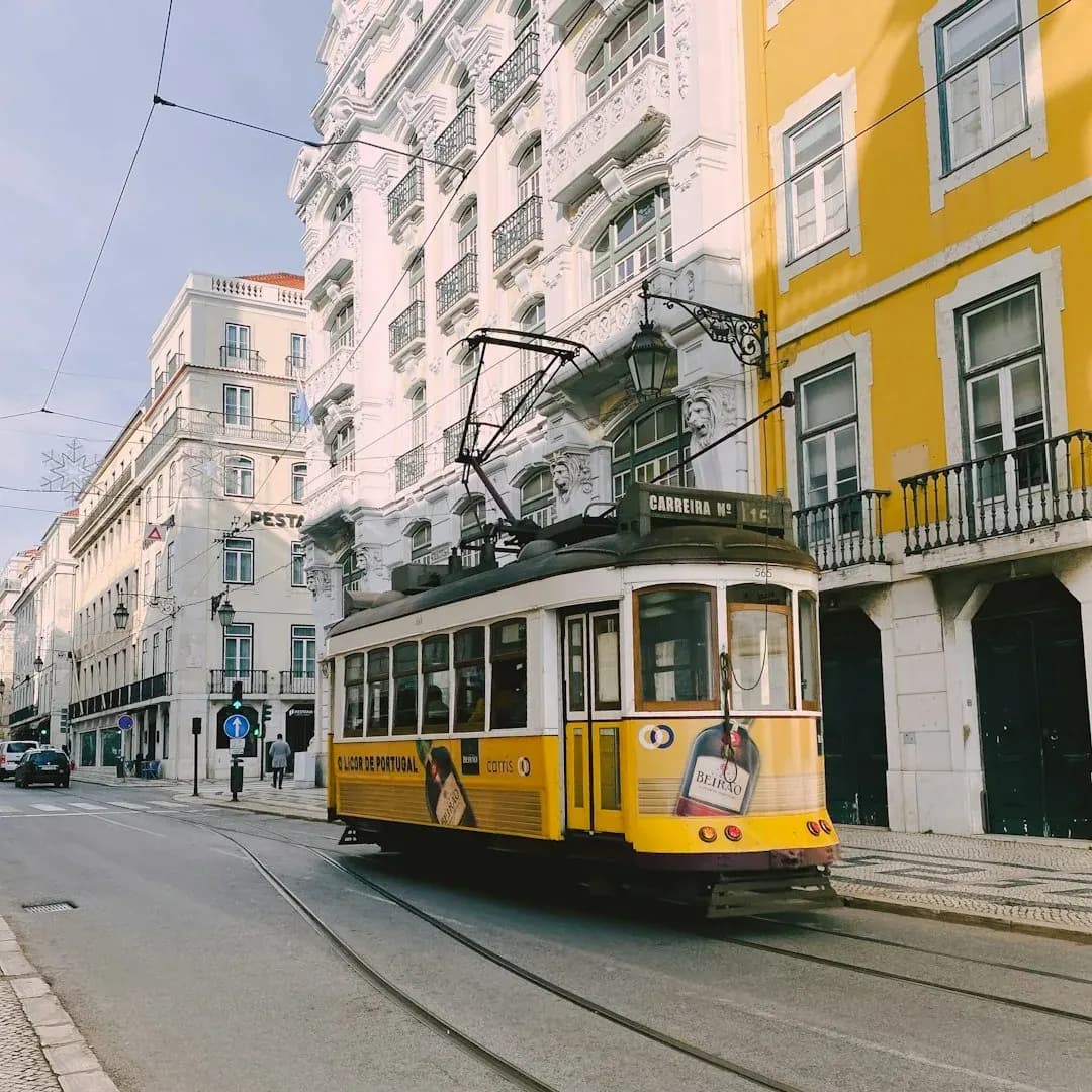 A warm, wide illustration of Lisbon at golden hour: the Praca do Comercio facing the Tagus estuary, the 25 de Abril bridge in the distance, terracotta rooftops stepping up the hills, under a clear amber sky.