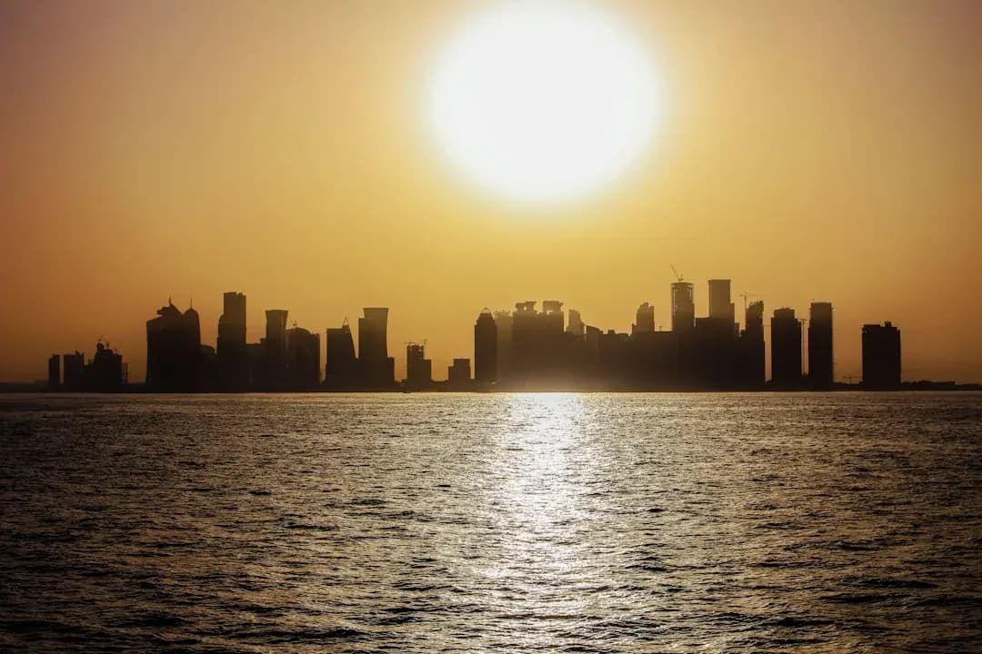 A wide warm illustration of Doha at golden hour: the West Bay skyline of glass towers along the Corniche, the curved Museum of Islamic Art on its island, and dhow boats on a calm blue bay in the foreground.