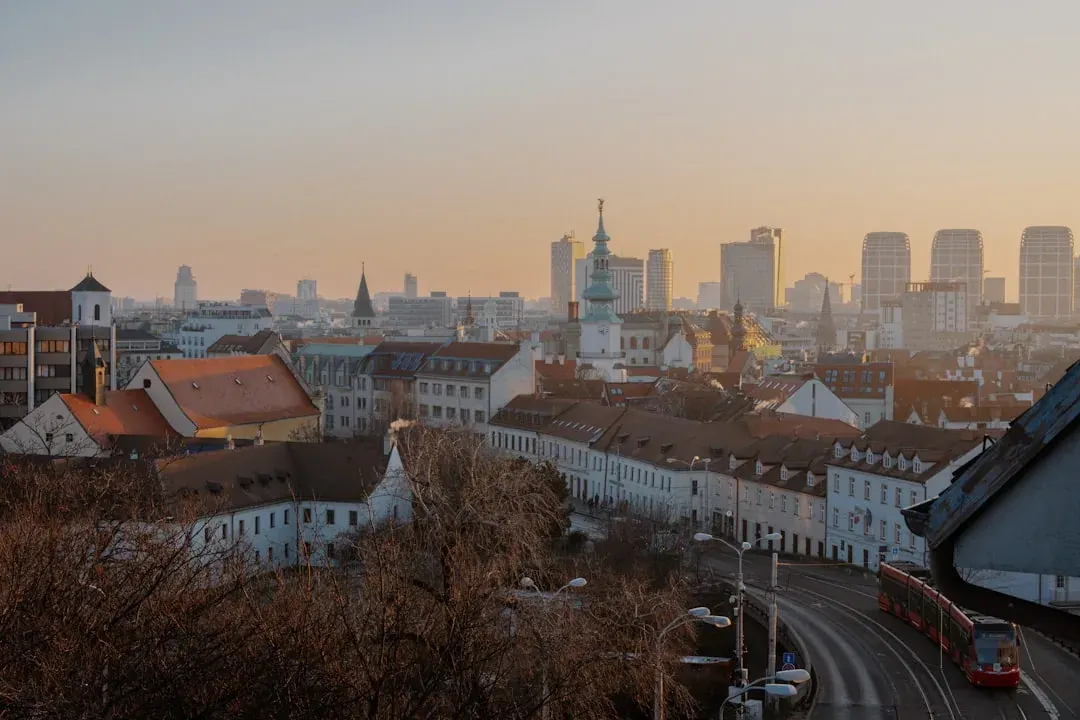 A wide warm illustration of Bratislava at golden hour: the castle on the hill above the Danube, the old town's pastel rooftops and St Martin's Cathedral spire, with the SNP Bridge tower catching the amber evening light.