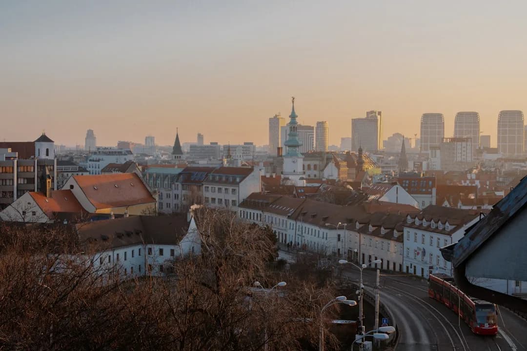 A wide warm illustration of Bratislava at golden hour: the castle on the hill above the Danube, the old town's pastel rooftops and St Martin's Cathedral spire, with the SNP Bridge tower catching the amber evening light.
