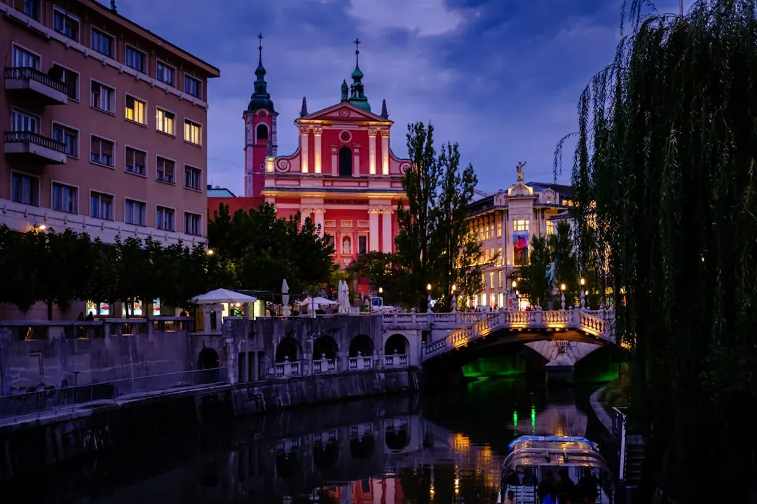 A wide warm illustration of Ljubljana at golden hour: the Ljubljanica river curving past the pastel facades of the old town, the Triple Bridge in view, and the castle hill rising green behind under an amber sky.