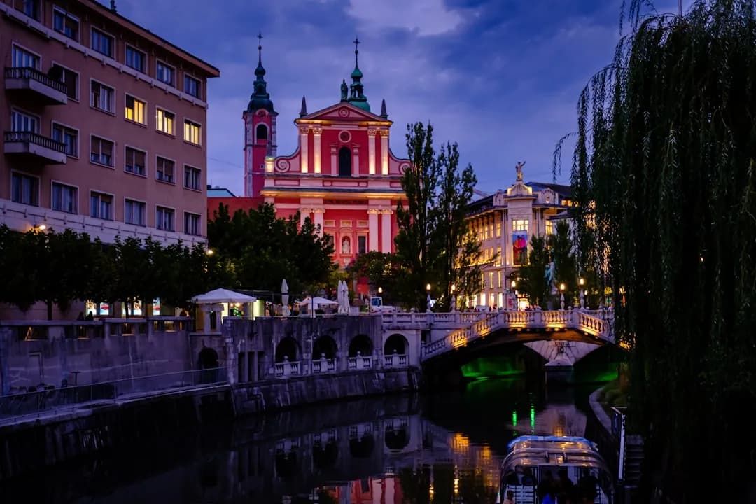 A wide warm illustration of Ljubljana at golden hour: the Ljubljanica river curving past the pastel facades of the old town, the Triple Bridge in view, and the castle hill rising green behind under an amber sky.