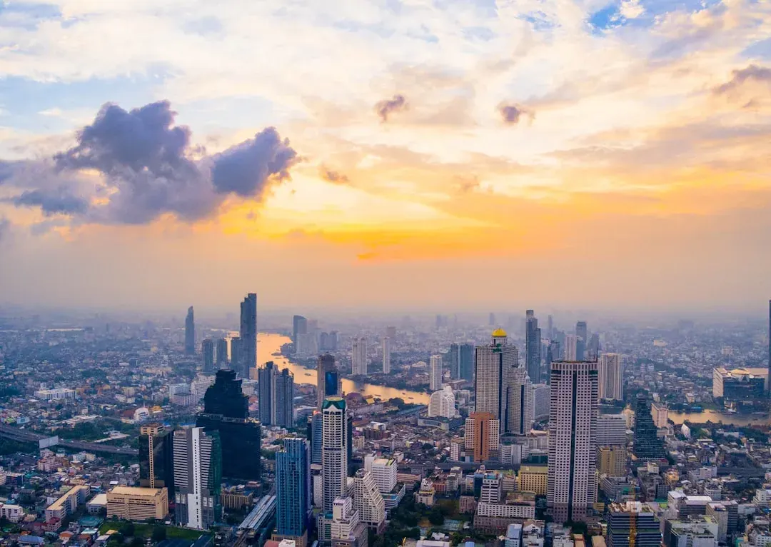 A warm wide illustration of Bangkok at golden hour, with the Chao Phraya river curving past riverside towers, longtail boats on the water, and the spires of Wat Arun catching amber light.