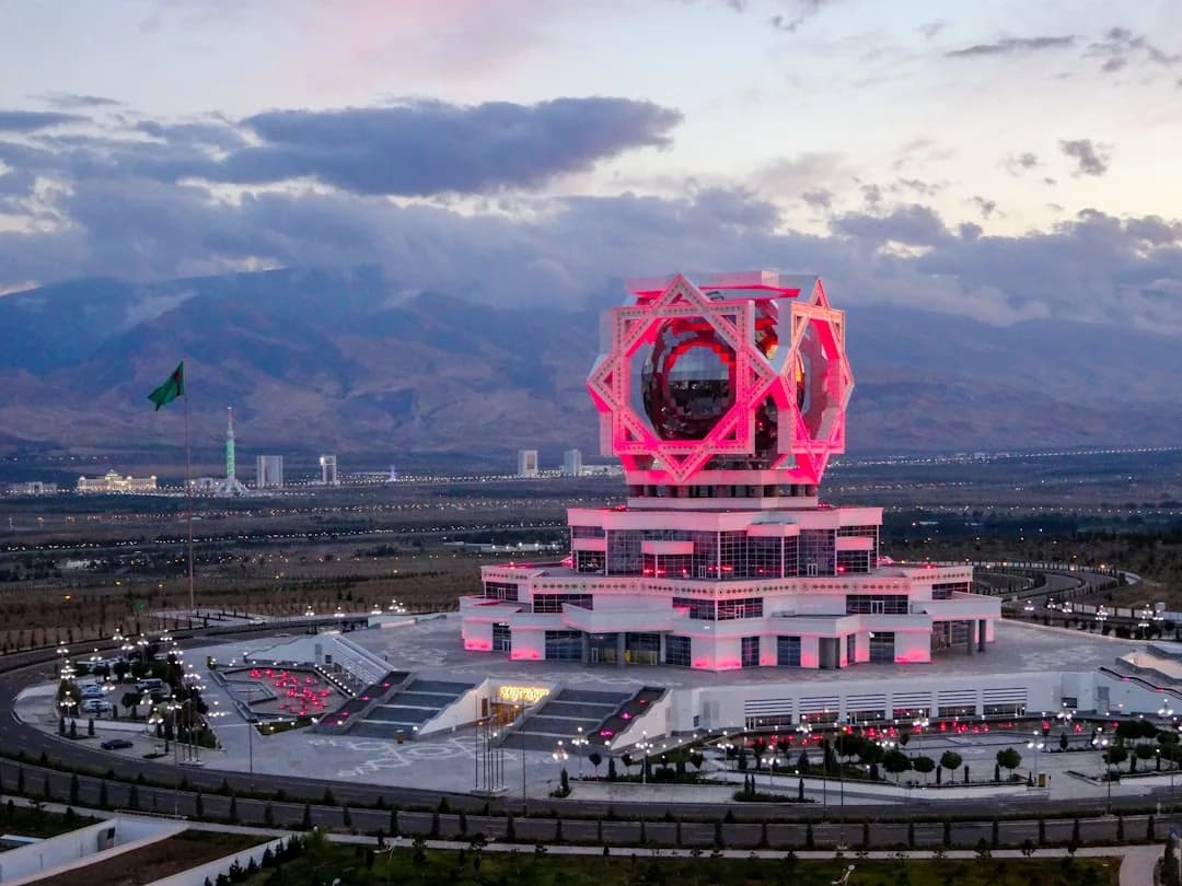 A freelance contractor working at a desk in Ashgabat, with the white marble cityscape and the Turkmenbashi World of Fairy Tales park visible through the window in warm afternoon light.