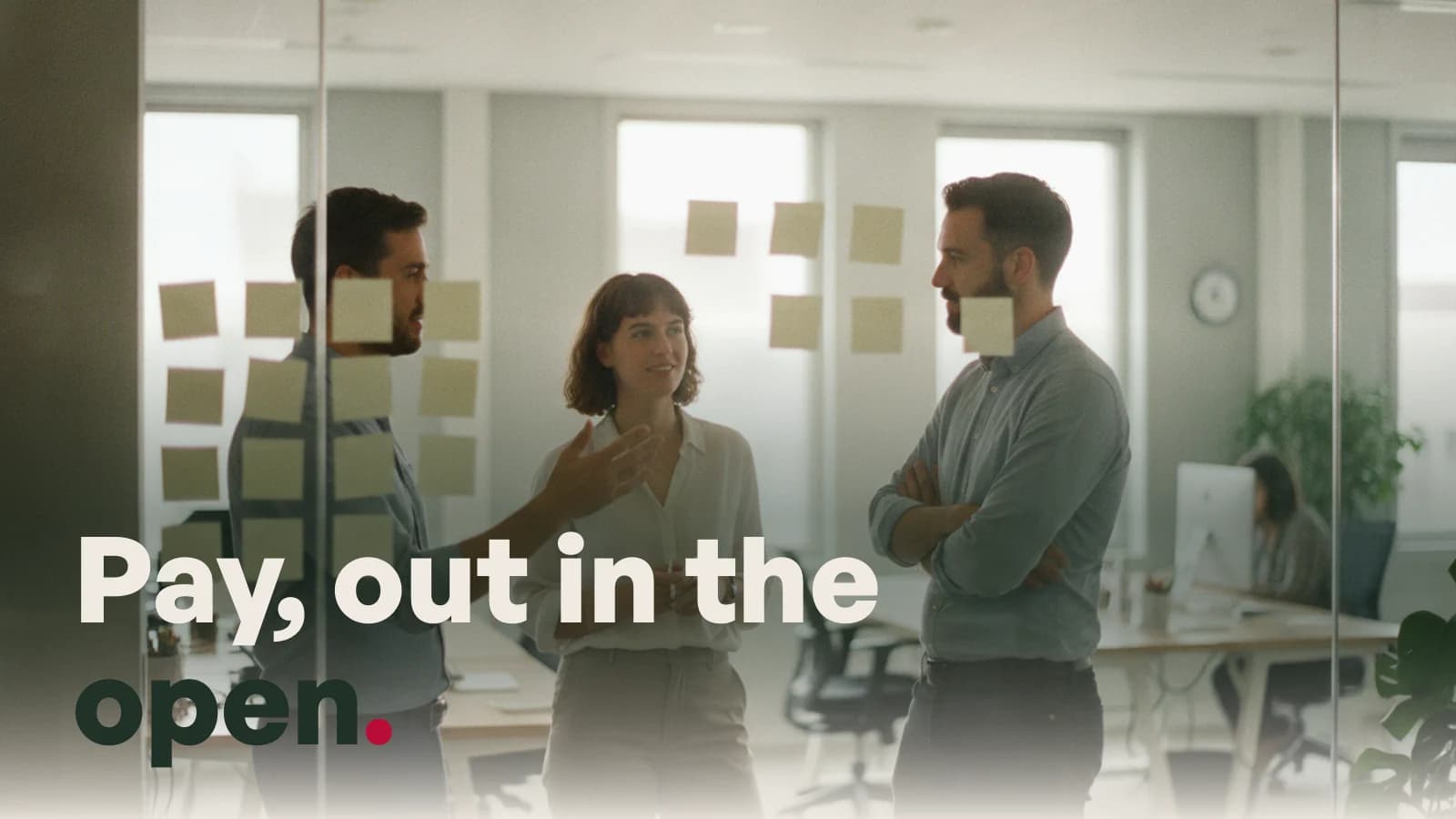 Three colleagues talking beside a glass office wall of sticky notes, captioned Pay, out in the open.