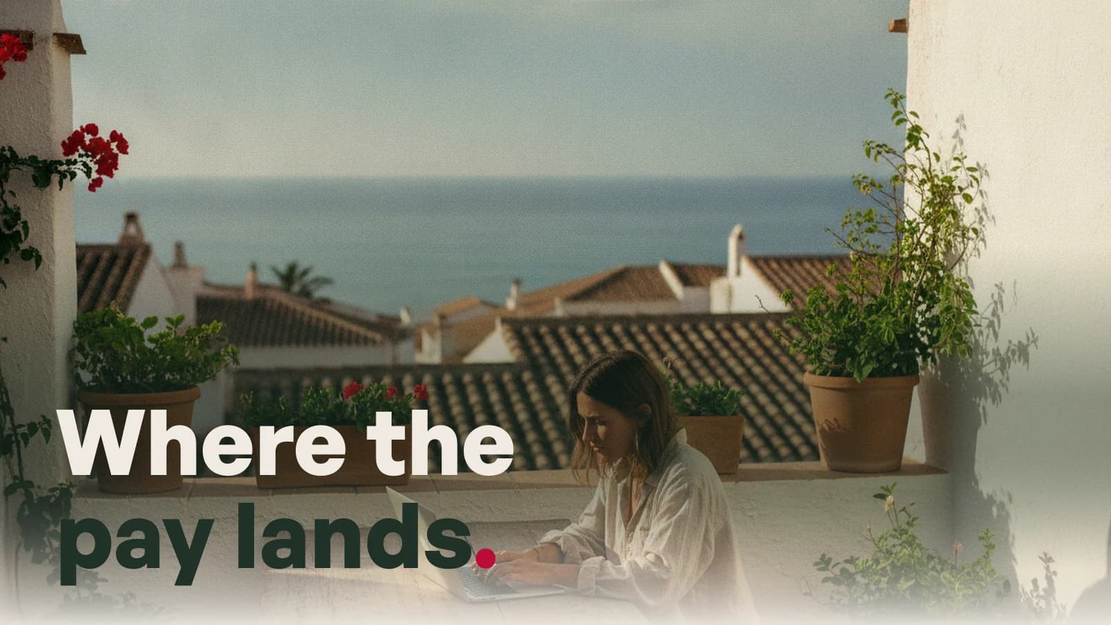 A woman working on a laptop on a shaded terrace in a Spanish coastal town, with the line Where the pay lands.