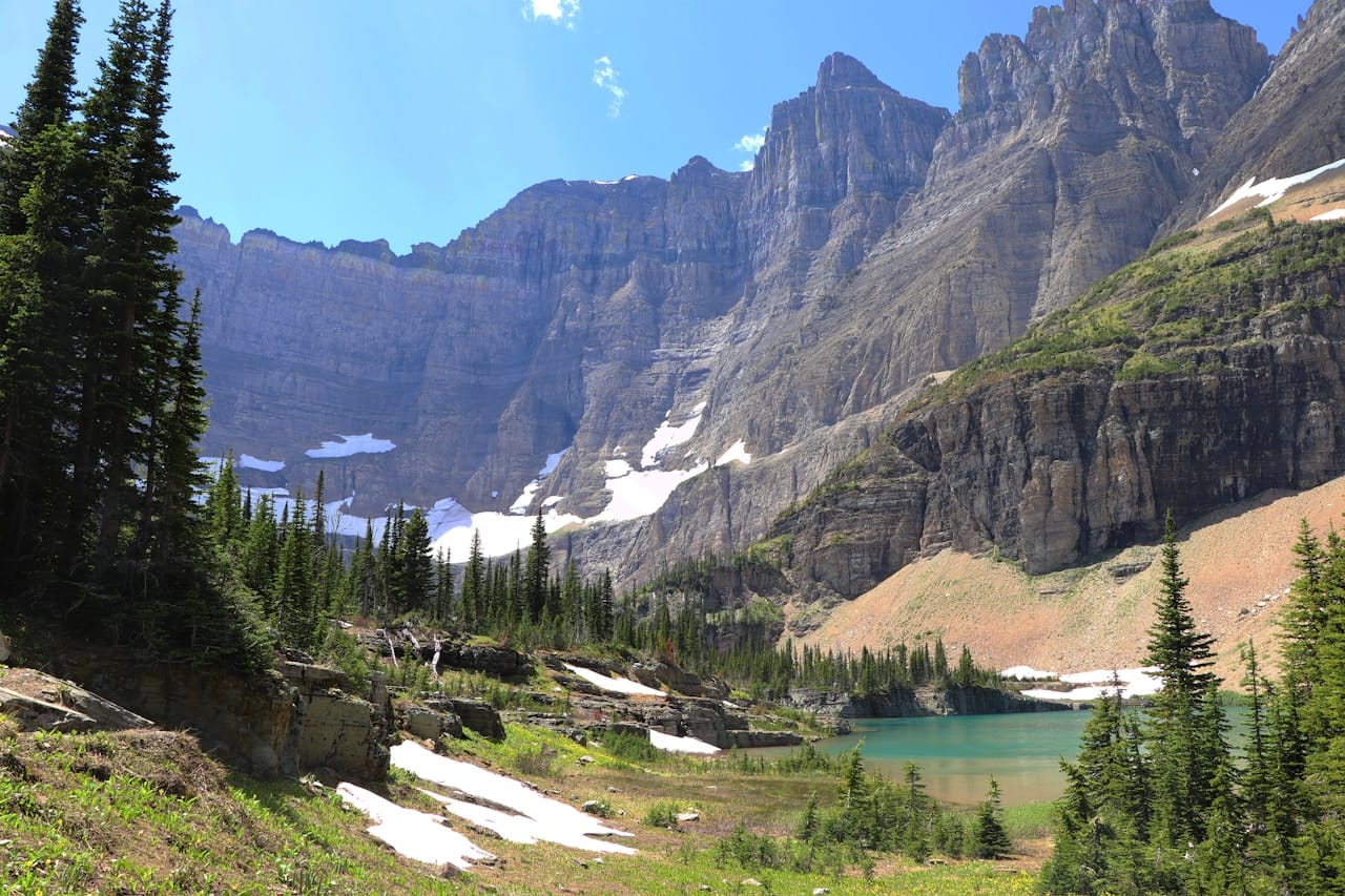 A sweeping panorama of Glacier National Park, Montana, with jagged peaks reflected in a clear mountain lake surrounded by pine forest.
