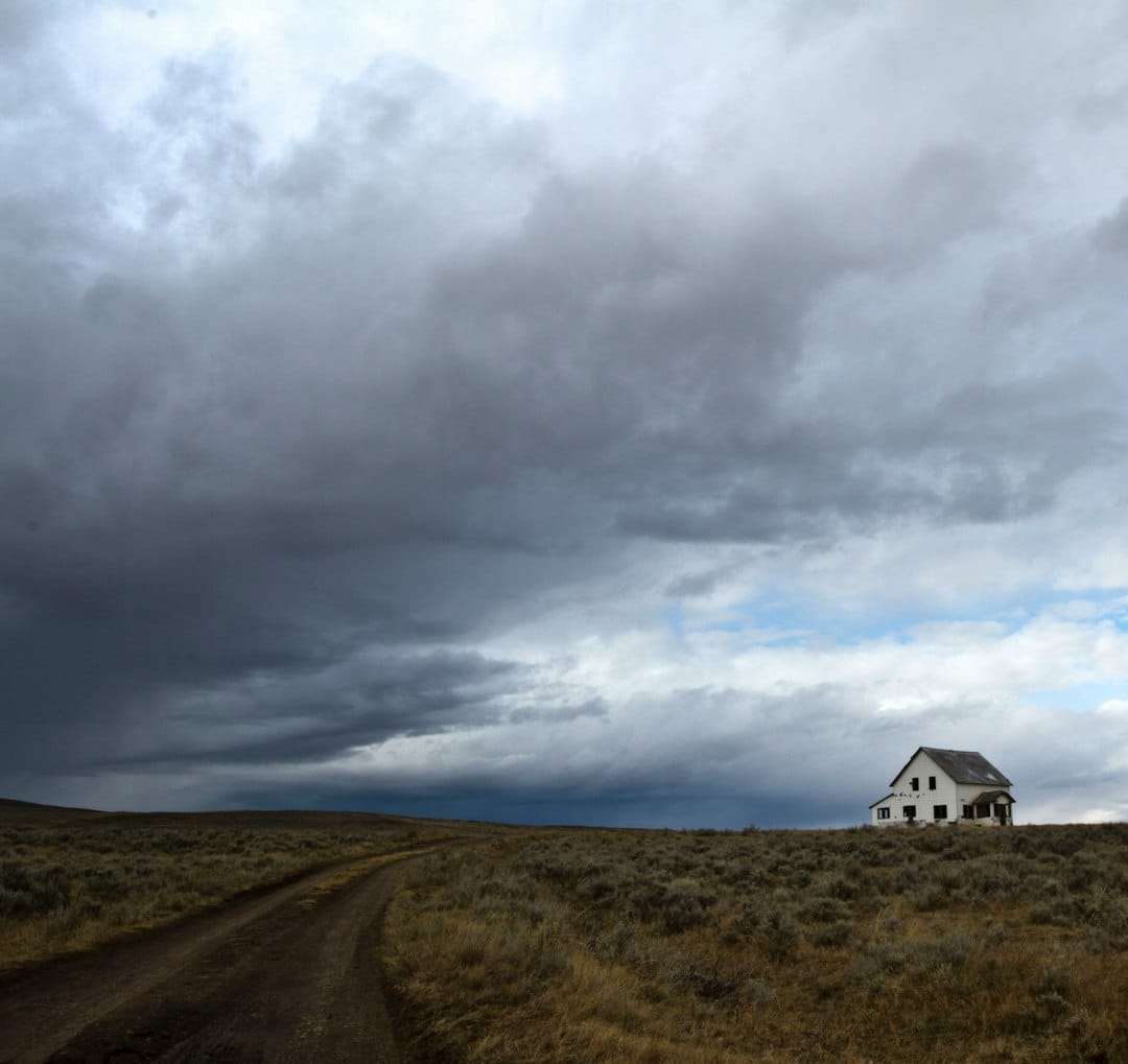 A wide-open Montana field under a dramatic big-sky cloudscape, with a farmhouse on the horizon.