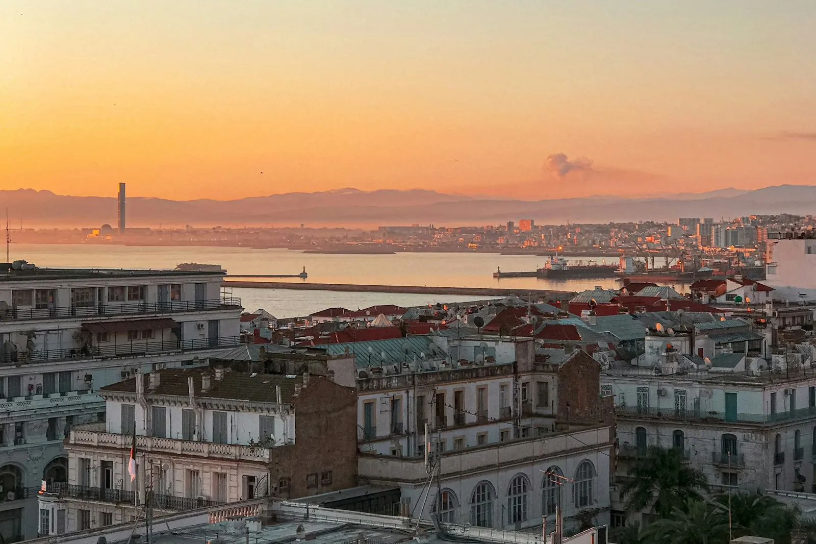 Rooftops and balconies of Algiers at sunset, with the bay and the distant coast glowing orange behind them.