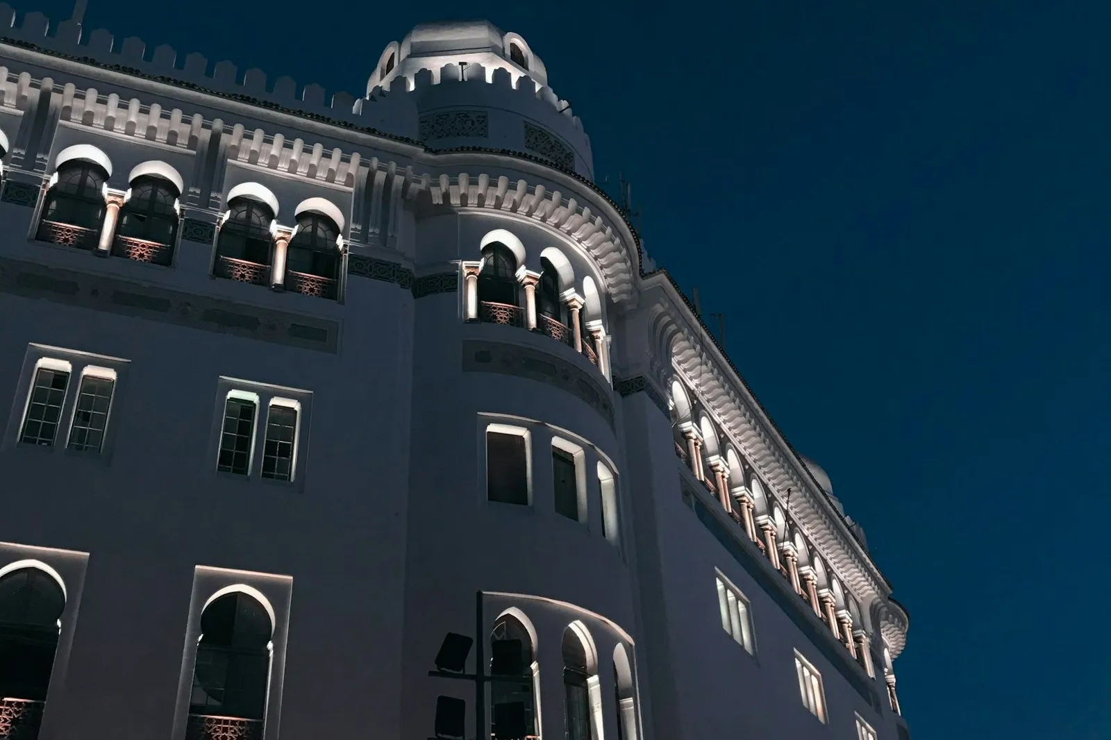 A white building lit at night in Algiers, with arched windows, a small round tower and a deep blue sky behind it.