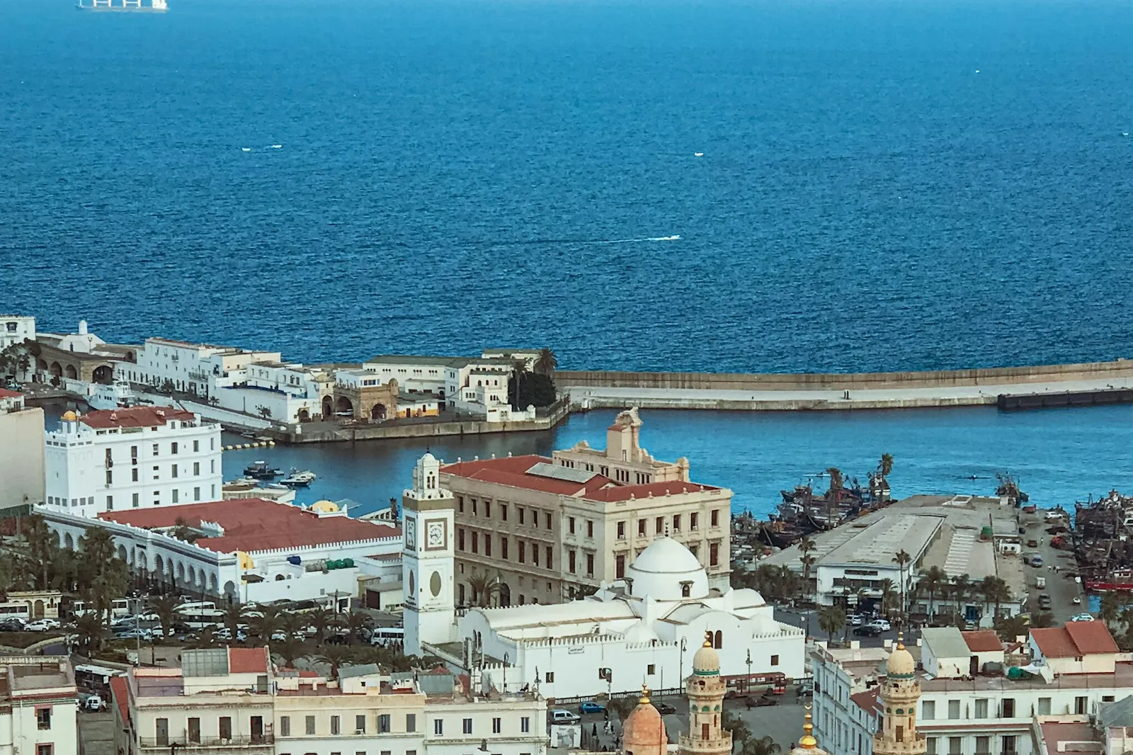 Algiers old town and harbour seen from above: white buildings with red roofs, a clock tower and a small dome beside the deep blue Mediterranean.