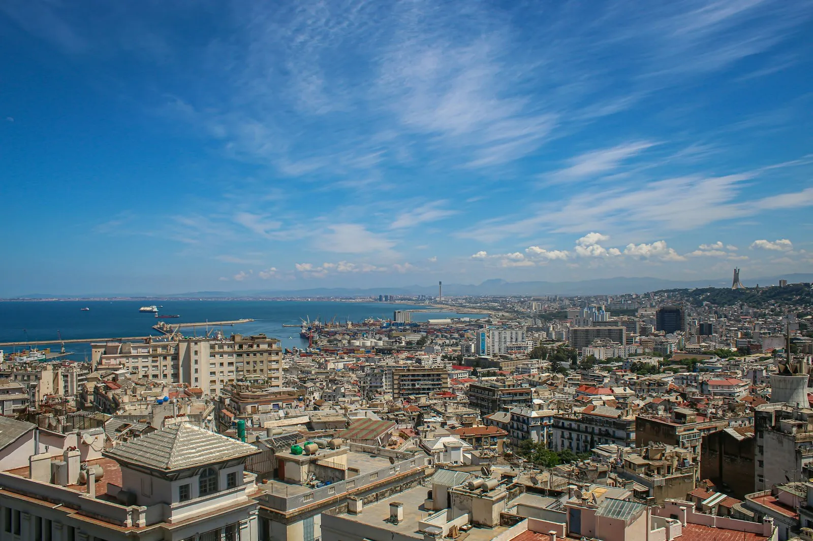 A view over Algiers, the capital of Algeria: dense white and cream buildings in the foreground, the bay and the harbour behind, under a blue sky with thin cloud.