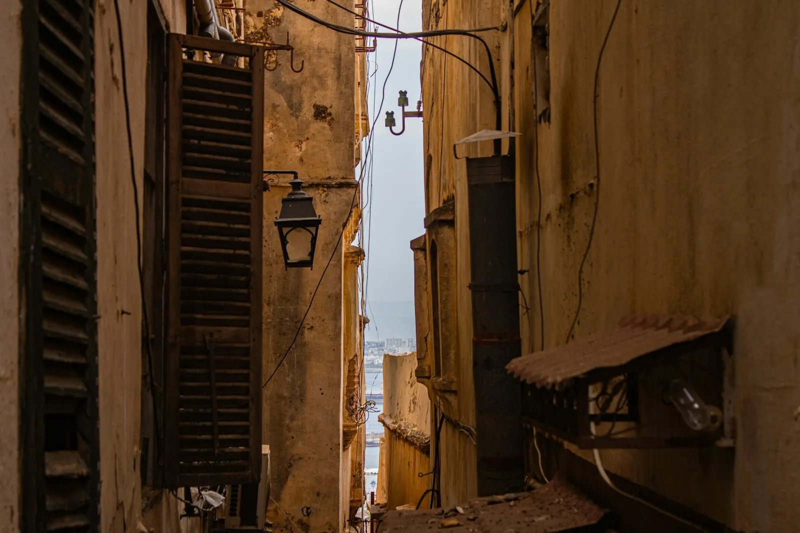 A narrow alley between weathered stone buildings in Algiers, with wooden shutters, a street lamp and a slice of sea at the end.