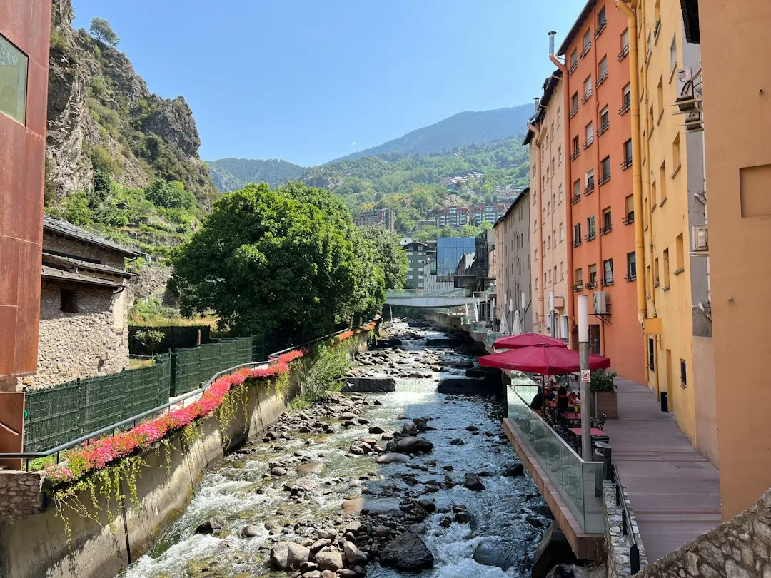A mountain stream running over rocks between a flowering railing and a row of orange and yellow apartment buildings in Escaldes-Engordany.